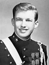 A black-and-white photograph of Donald Trump as a teenager, smiling and wearing a dark pseudo-military uniform with various badges and a light-colored stripe crossing his right shoulder. This image was taken while Trump was in the New York Military Academy in 1964.