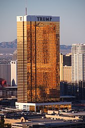 A tall rectangular-shaped tower in Las Vegas with exterior windows reflecting a golden hue. It is a sunny day and the building is higher than many of the surrounding buildings, also towers. There are mountains in the background. This tower is called the Trump International Hotel Las Vegas.